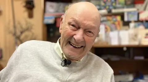 Malcolm Metcalf smiles at the camera. He is wearing a pale grey sweatshirt and a pale lemon shirt.