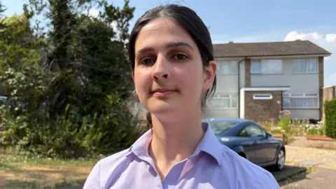 Hannah standing on a residential street in front of a row of semi-detached houses. She is centred in the frame, wearing a light purple button-up shirt and looking towards the camera. Parked cars, including a blue coupe and a white SUV, are visible on driveways behind them. Trees and greenery line the left side of the scene, with bright daylight and a blue sky overhead.