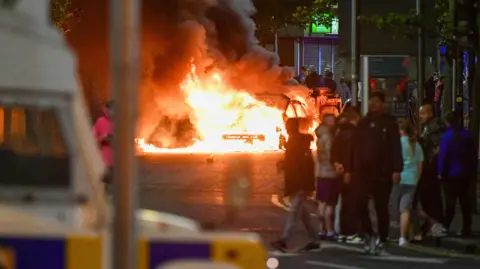 Pacemaker Car burning in Belfast with crowd stood observing