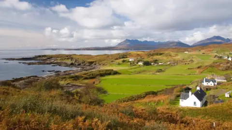 White-walled houses scattered across a coastal landscape of fields and areas of rugged shoreline. In the distance are Skye's Cuillin mountains.