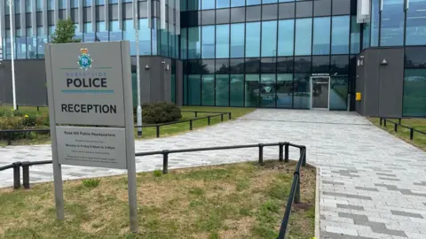 An exterior view of a multi-storey glass fronted office building with a grey paved pathway towards a sliding door. In the foreground is a sign on a small patch of grass reading 'Merseyside Police reception'. 