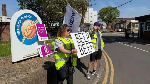 A man and a woman in high visibility jackets hold up a flag and a placard next to a road. Behind them is a sign for Weetabix Food Company.