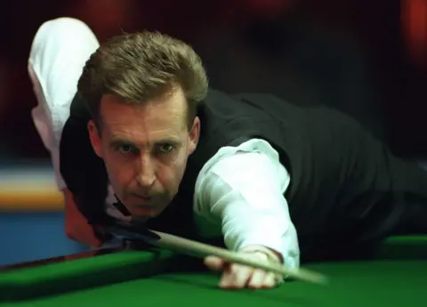 Terry Griffiths bent over the snooker table, cue resting on the bridge between his thumb and index finger on his right hand as he prepares to strike the white ball. He is wearing snooker attire, with a white long sleeve shirt and black vest.