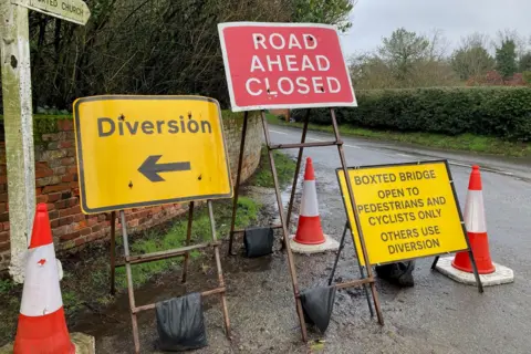 James Hore/BBC A red sign reads ROAD AHEAD CLOSED. And it is flanked by two yellow road signs. A country road can be seen behind. There are hedges and trees.