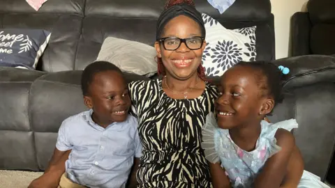 Perpetual pinch her boy Osinachi and her girl Sochika. They are sitting connected nan level astatine location successful beforehand of a sofa. Perpetual has her arms astir nan twins. They are each smiling and looking happy.