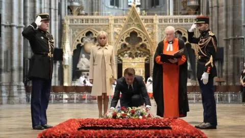 PA Media Emmanuel Macron, centre, kneels down to place a colourful wreath at a plaque on the ground inside Westminster Abbey, which is bordered by poppies. Brigitte stands solemnly to the left, and the sub-Dean of Westminster Abbey, Reverend David Stanton stands on the right. They are flanked by two soldiers in black uniform standing at salute.