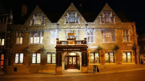 The outside of a hotel, all lit up. It has an animal above a canopy and there are lots of windows in the old stone building. There is a large door at the front, in the middle of the building. 