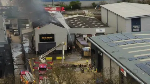 Paul Wood A view from above of the industrial estate, with fire engines parked and one extended ladder. One building is on fire - it has the words Neo Bros Car Parts on the front. Crews spray water over it from a yellow hose.