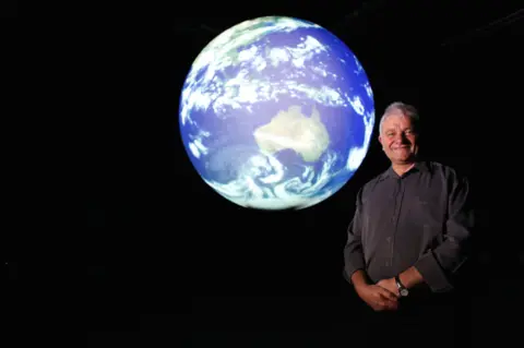 BBC A man stands in a dark room before a large glowing globe of the Earth, prominently showing Australia, with his face and folded hands softly lit by the planet’s light
