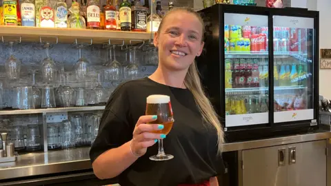 Henry Godfrey-Evans/BBC Bartender Deanne Follett holding a pint of beer in her right hand and smiling. She is standing behind the bar, with glasses, bottles and drinks all behind her.