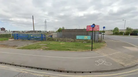 View of the derelict land that will become the car park there is a brick electricity substation in the foreground and the site is surrounded by metal fencing with a pavement and cycle path in front of it and a road on the right hand side 