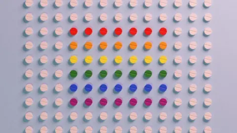 Getty Images Collums of white pills sit on a blue backround. There are 10 rows, with 13 pills in each row. In the centre of these pills there are muti-coloured ones. Each row is a different colour, from top to bottom they're red, orange, yellow, green, blue and purple.
