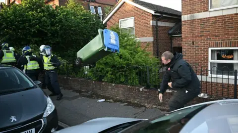 Getty Images A man in a residential street throws a wheelie bin in the direction of a group of police officers dressed in riot gear. 