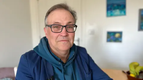 A head and shoulder shot of Philip, who has grey hair and wears a pair of spectacles. He is wearing a blue hoodie with a blue cardigan on top of it. In the background you can see various paintings and some bananas in a basket.