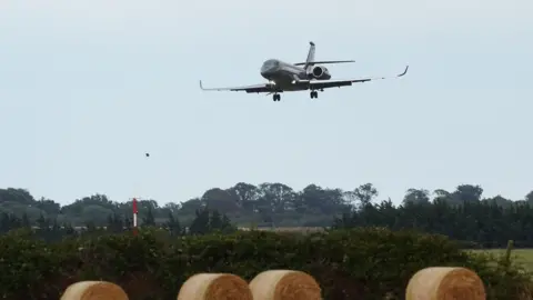 PA Media A dark grey plane comes into land near a field of hay bales and green trees, the sky behind the aircraft is blue.