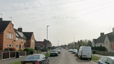 A Google streetview image of a long residential road with houses on both sides of the road. Cars and a van can also be seen parked on both sides.