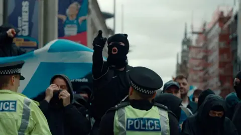 Person in balaclava pointing while shouting as a police officer watches