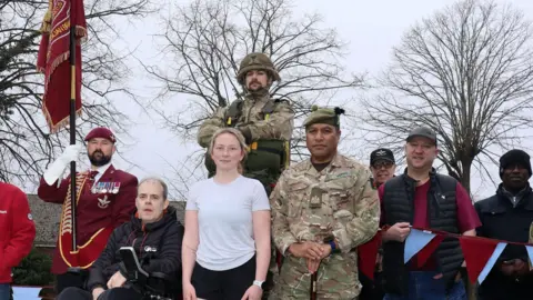 L/Cpl Mackenzie Delaney in white T-Shirt and black shorts is stood with the representatives of the garrison in uniform and other local dignitaries. They are outdoors with trees behind them. A flag bearer in a burgundy coloured uniform holds a garrison standard. 