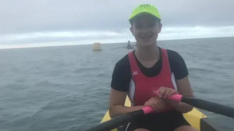 Jersey Rowing Club Ryanna is sitting in a small rowing boat holds an oar with pink grips, wearing a bright green cap and red athletic vest, with open water and a buoy visible under a cloudy sky.