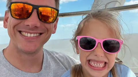 Matt Ridout is wearing reflective sunglasses and a grey T-shirt and is grinning at the camera. He has his arm round his daughter, Amelia, who had long blonde hair tied back and wore pink sunglasses. She is smilling so you can see both her upper and lower teeth. They are sitting, with the sea in the background.