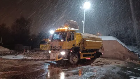 Staffordshire County Council A yellow gritter can be seen as snow falls in front of it. The gritter is a lorry with a load of brown salt on the back. Behind it is a further pile of salt on the ground.