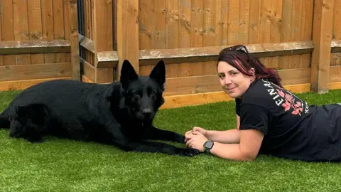 A woman and a black retired police dog - called Marco - sat on astroturf. 