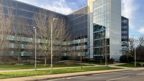 Nicola Haseler/BBC A modern multi-storey car park with glass-fronted stairs section. Trees that have lost their leaves are on the grass verges in front of it. White emergency service tape runs around the building.