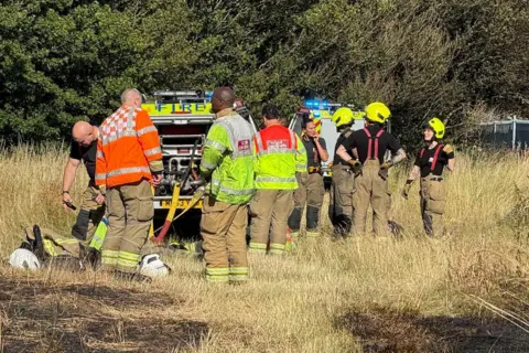 Craig Fallon A group of eight firefighters in uniform stand on parkland, some of which is burnt, near two fire vehicles.