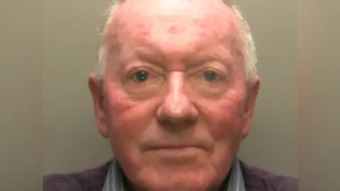 Police mugshot of Ronald Short. He has short grey hair, and is wearing a purple jumper and grey shirt. He is looking directly at the camera 