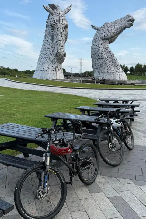 PATRICK MORAN Two mountain bikes leaning against empty picnic tables with the giant Kelpies sculptures in the background