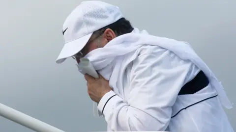 PA Media Megrahi climbing the steps of a plane at Glasgow Airport. He wearing a white tracksuit, white Nike baseball cap, and is holding a white scarf around his face.