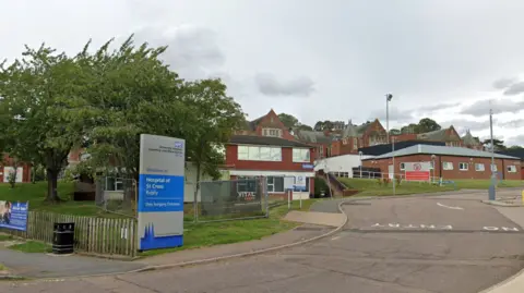Google The front of the Hospital of St Cross with a blue and silver NHS sign and brick buildings in the background