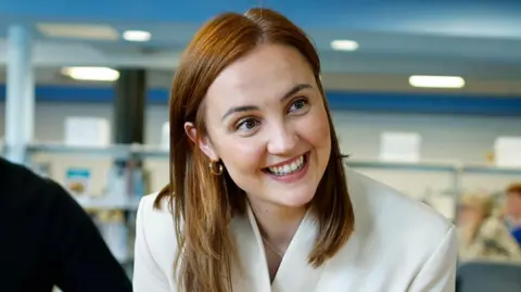 Getty Images Head and shoulders shot of a smiling Mairi McAllan in a classroom setting. She is wearing a white jacked and has long brown hair.