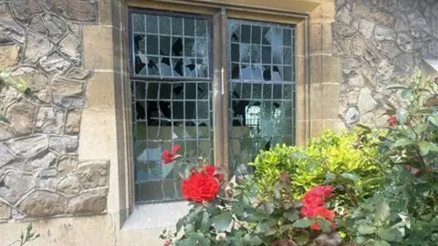 Southend City Council A damaged window with broken glass panes on a stone building, partially obscured by a vibrant red rose bush.