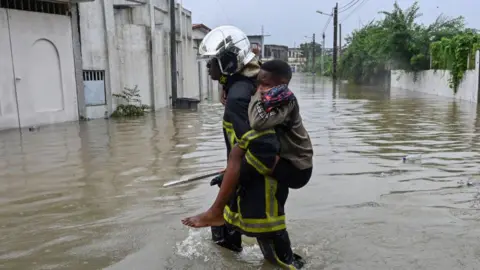 Issouf Sanogo/AFP A firefighter carries a child as he walks in flood water in Cocody Angre in Abidjan on June 14, 2024 after torrential rains. Flooding and landslips have killed five people in Abidjan, Ivory Coast's biggest city, after heavy downpours, the fire service said on Friday. Roads were cut off as the rains fell on Thursday afternoon in most areas of the city with a population of six million.