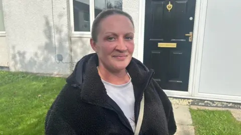 A woman stands in a garden in front of a black front door, weearing a bobbly dark fleece and a white t-shirt. Her hair is tied back and she is smiling while looking directly at the camera.