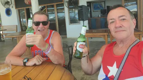 Metropolitan Police Mr Longworth, left, and Mr Alfonso, right, smile at the camera. They are both holding green bottles of Stella and wearing red vest tops with blue and white graphics on them. They are sitting at what appears to be an outdoor bar or restaurant, overlooking a view.