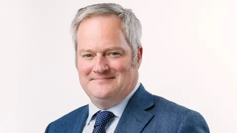 Sussex Partnership Dr Oliver Dale. He has grey hair and is wearing a light coloured shirt, a spotted navy tie and a blue blazer 