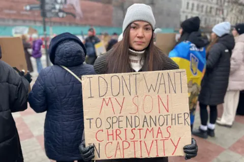 A woman is seen holding a cardboard sign which says "I don't want my son to spend another Christmas in captivity".