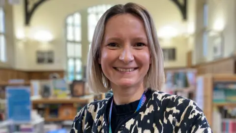 The picture shows Sarah Herlinger smiling at the camera. She is standing in the school's library