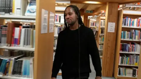 Jason Arday, dressed in black, walks through a library