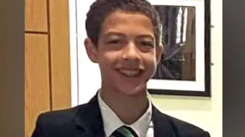 Noah Donohoe, with short brown hair, smiling at the camera. He is wearing a white shirt, green and black tie and a black blazer.