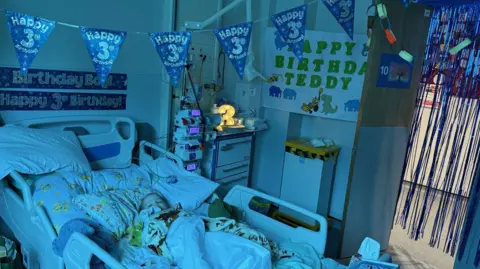 Sarah Sloman A young biy in a hospital bed, wrapped in cartoon-print blankets. Above him is blue bunting which says "Happy 3rd Birthday". There is a light in the shape of a 3 on the bedside table, and a sign on the wall which reads "happy birthday teddy" as well as blue sparkly fringing framing the door.