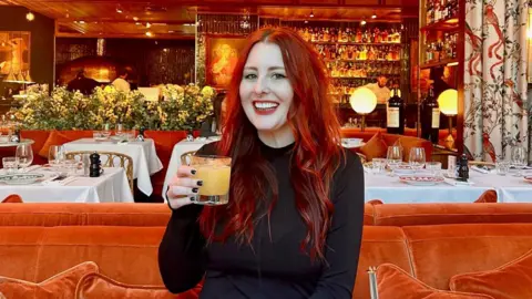 A woman with long dyed red hair and wearing a black long-sleeved top is sitting in a restaurant on an orange sofa. She is holding a light orange drink in a glass and smiling at the camera.