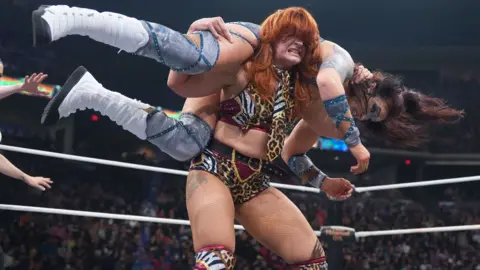 AEW Jamie Hayter - a woman with red hair, wearing a red cropped top and red and gold shorts, holding an opponent on her shoulders in a wrestling ring