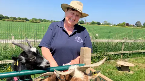 Gillian Henshaw wearing a branded navy polo t-shirt and a beige sunhat. She is stood on a farm feeding goats.