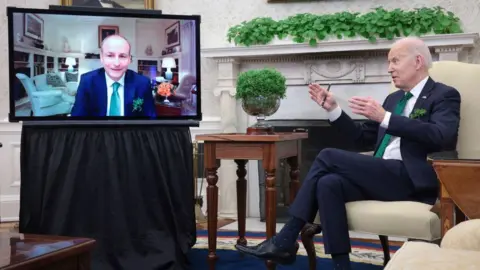 Getty Images Micheál Martin se muestra en una pantalla de televisión en la Casa Blanca durante una videollamada con el ex presidente de los Estados Unidos, Joe Biden, en marzo de 2022. Ambos hombres usan trajes azul marino, camisas blancas y corbatas verdes. Un tazón de Shamrock está en una mesa frente a Biden. 