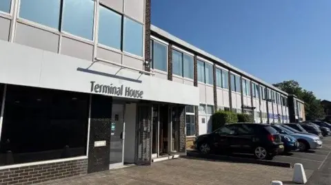 A grey-cladded office building with a row of cars parked in front. The building bears the sign 'Terminal House'.