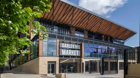 A canopied market building, heavily glass fronted. A set of black metal gates lead into what looks like a courtyard area.