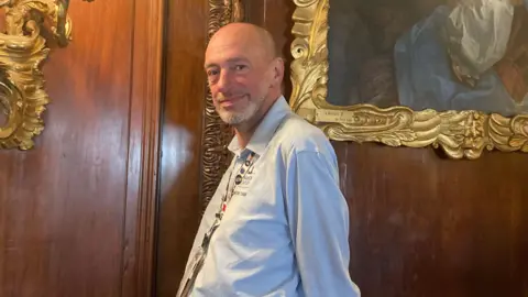BBC/Richard Williams A man with a shaved head and pale shirt, with a lanyard round his neck. He is standing in front of wooden panelling and there are portraits in gilt frames on the walls.